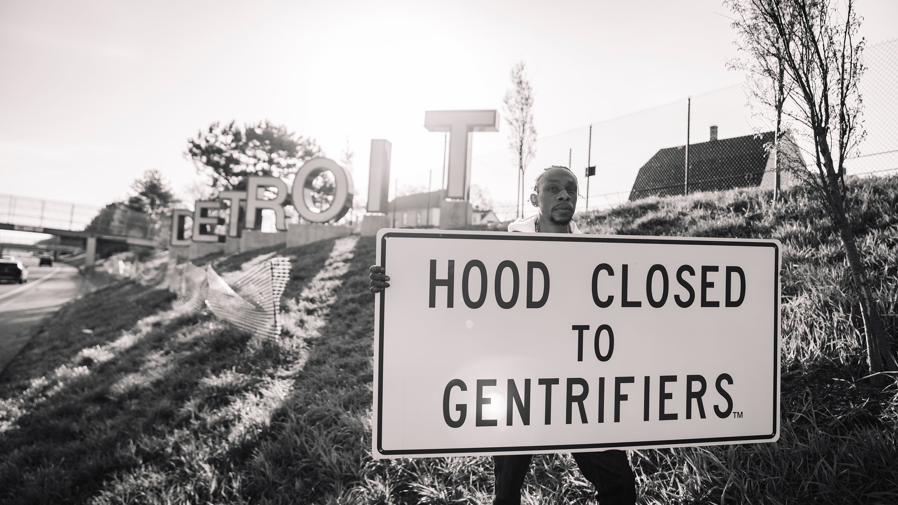Person holding a sign that reads 'Hood Closed to Gentrifiers' with a large 'DETROIT' sign in the background.